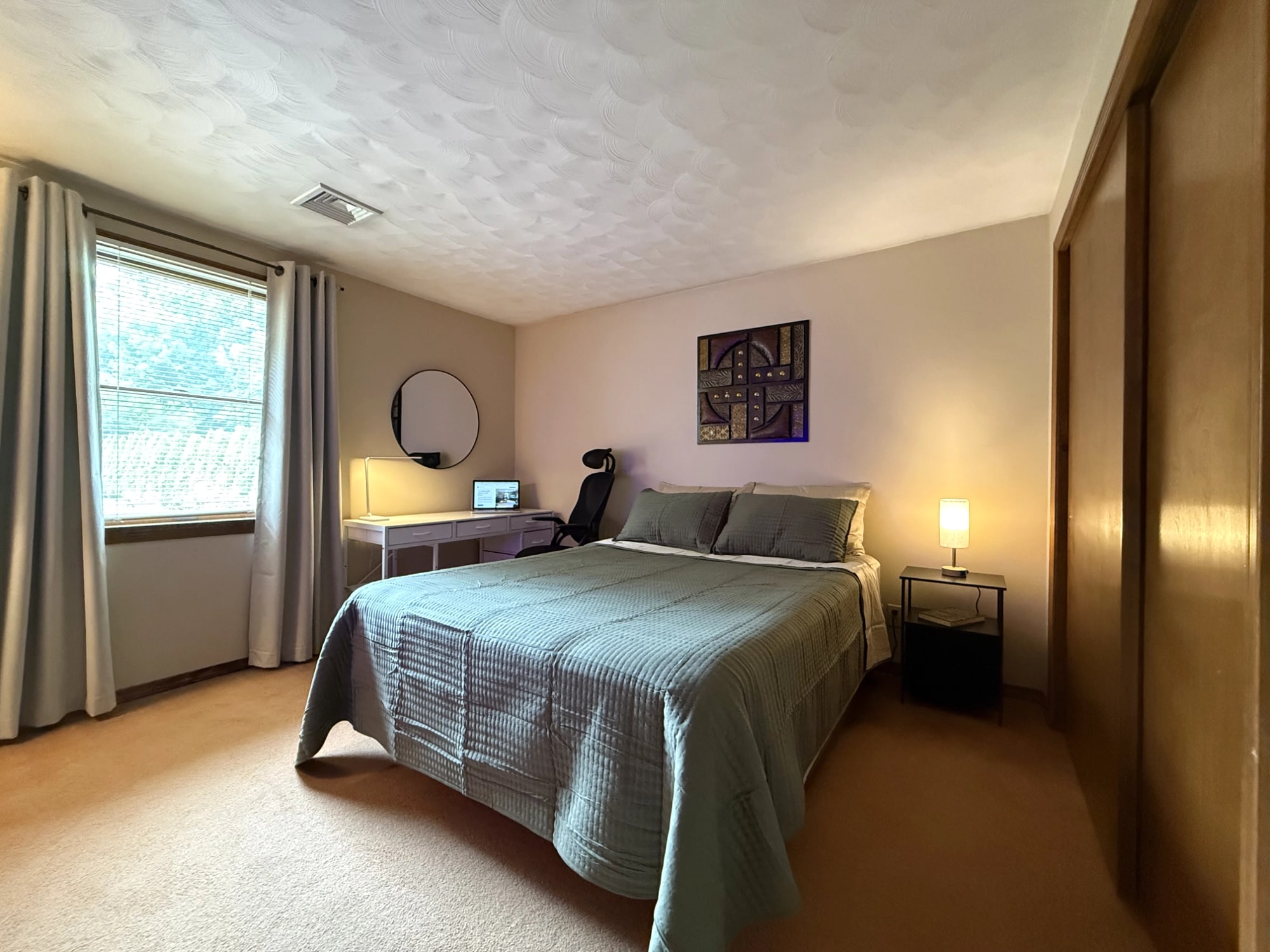 Furnished queen room with bed, window, desk and bedside lighting